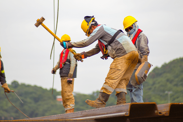 Mehrere Bauarbeiter auf einem Dach, die einen Hammer hochheben und dabei Sicherheitsschuhe für Bauarbeiter tragen.