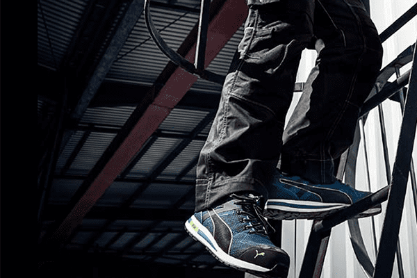 Die Beine eines Logistikers, stehend auf einer Leiter. Trägt blau-schwarze Sicherheitsschuhe