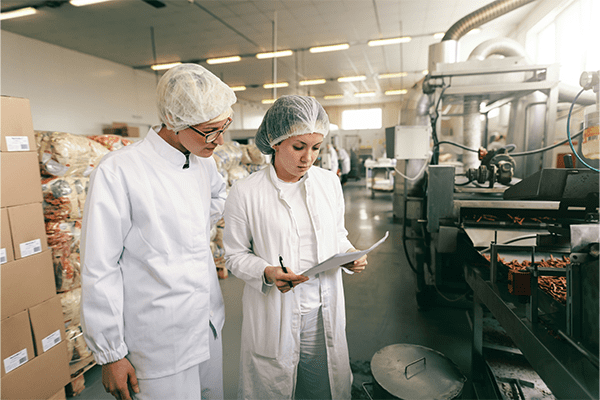 Lebensmitteltechnologinnen arbeiten in einer Fabrik. Sind weiss gekleidet mit Haarhaube.